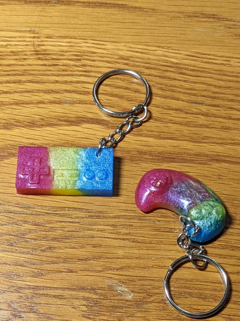 May include: Two keychains with silver rings and chains. One is a rectangular shape with a controller design in pink, yellow, and blue. The other is a curved shape in pink, purple, green, and blue.