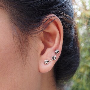 May include: A silver ear cuff with four small rose-shaped charms. The cuff is worn on the upper part of the ear, curving up and away from the earlobe.