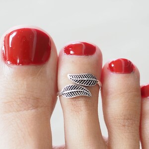 Op de afbeelding: Een zilveren teenring met een bladerenontwerp. De ring zit op een teen van een persoon met rode nagellak.