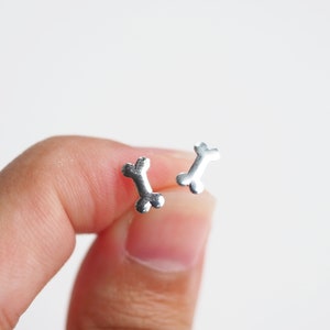 May include: A pair of small, silver-colored bone-shaped stud earrings. The earrings are held against a white background. The earrings are a simple, minimalist design.