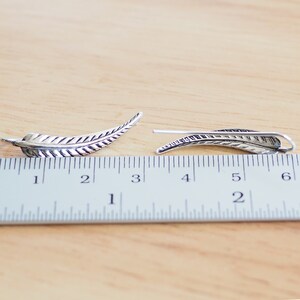 May include: Two silver feather earrings, each measuring approximately 2 inches in length, are displayed on a wooden surface with a ruler for scale.