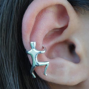 May include: Silver ear cuff in the shape of a person climbing. The cuff is worn on the upper part of the ear.