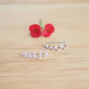May include: Two silver ear cuffs with a chain of connected circles. The ear cuffs are on a light brown wooden surface.