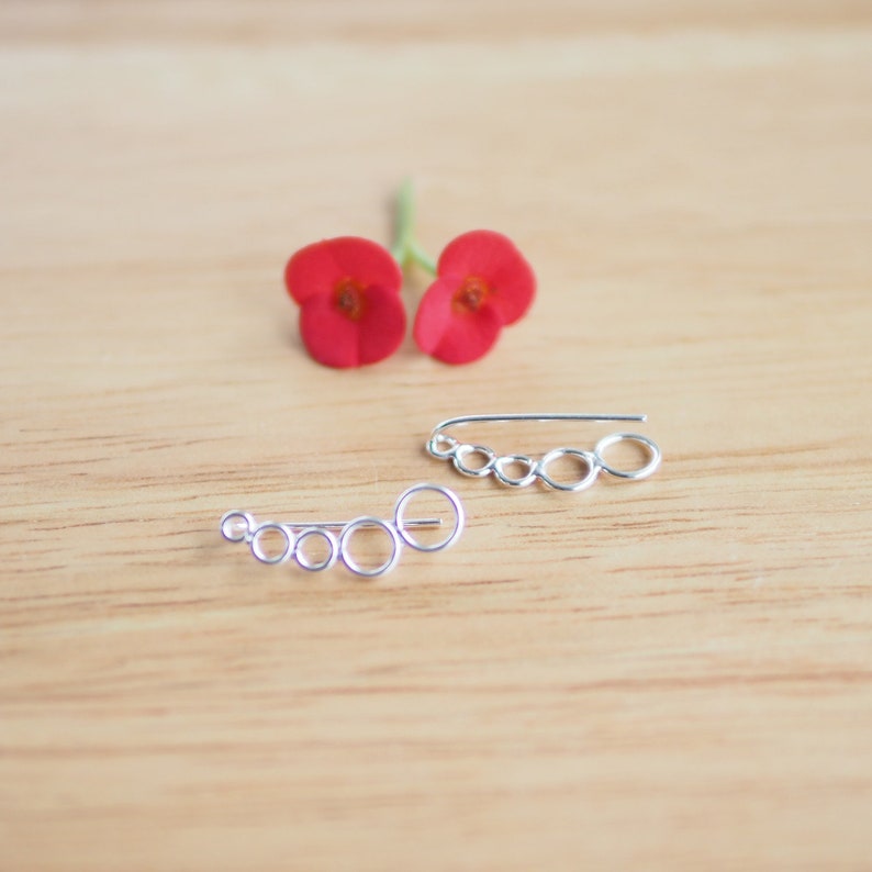 May include: Two silver ear cuffs with a chain of connected circles. The ear cuffs are on a light brown wooden surface.
