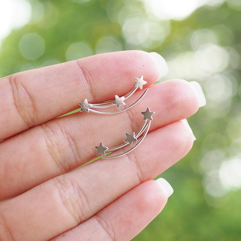 May include: A pair of silver ear cuffs with five star-shaped charms arranged in a curved line, resembling a shooting star.