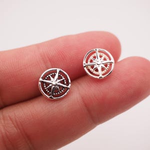 Könnte beinhalten: Ein Paar kleiner, silberfarbener Kompassrosen-Ohrstecker. Jeder Ohrring hat ein detailliertes Kompassdesign in einem kreisförmigen Rahmen. Die Ohrringe werden vor einem hellen Hintergrund gezeigt.