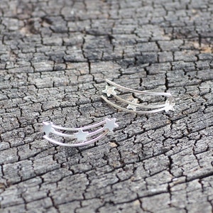May include: Two silver ear cuffs with a curved shape and a design of three stars in a row. The ear cuffs are on a wooden surface.