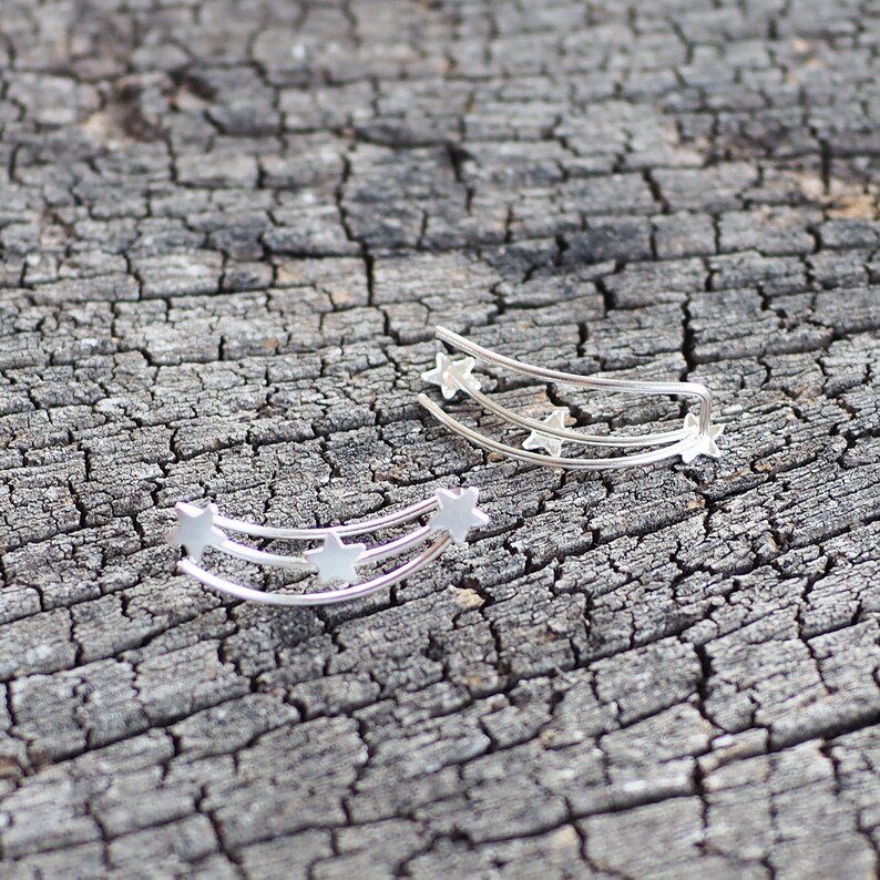 May include: Two silver ear cuffs with a curved shape and a design of three stars in a row. The ear cuffs are on a wooden surface.