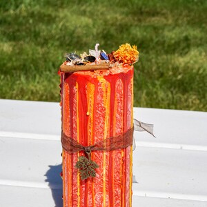 May include: A red and orange pillar candle with a decorative brown ribbon and a small leaf charm. The candle is decorated with drips of wax and has a floral topper.