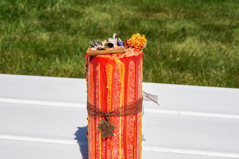 May include: A red and orange pillar candle with a decorative brown ribbon and a small leaf charm. The candle is decorated with drips of wax and has a floral topper.