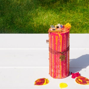 May include: A red and yellow striped candle with a brown ribbon and a small leaf charm. The candle is decorated with dried flowers and herbs.