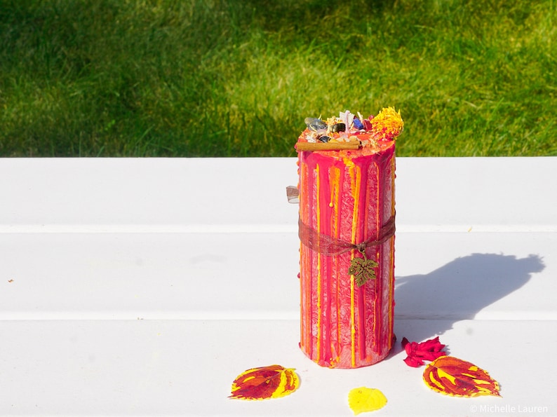 May include: A red and yellow striped candle with a brown ribbon and a small leaf charm. The candle is decorated with dried flowers and herbs.