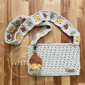 May include: A white crocheted bag with a long strap featuring a repeating pattern of yellow, green, and brown squares. The bag has a small tassel charm and a label that reads "The Cozy Dragonfly".
