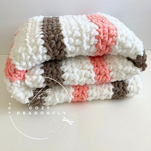 May include: A crocheted blanket with stripes of white, brown, and pink yarn. The blanket is folded in thirds and the text "The Cozy Dragonfly PDF Pattern" is visible on the bottom left corner of the image.