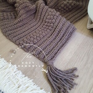 Puede incluir: Una manta de crochet marrón con un borla. La manta tiene un patrón texturizado y está hecha con un hilo suave. El texto "The Cozy Dragonfly Pattern" es visible en la imagen.