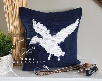 May include: A square navy blue crochet pillow featuring a large white silhouette of a bird in flight. The text The Cozy Dragonfly is printed in white on the front. The pillow is displayed alongside a braided cord and a small whistle.