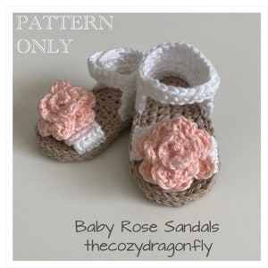 K&ouml;nnte beinhalten: Ein Paar geh&auml;kelte Babysandalen mit einer rosa Blumenverzierung. Die Sandalen sind beige und wei&szlig; mit einem wei&szlig;en Riemen. Der Text "Baby Rose Sandals thecozydragonfly" ist unter den Sandalen gedruckt.