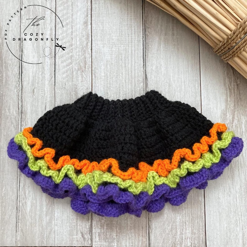 May include: A black crochet skirt with a ruffled hem in purple, green, and orange. The Cozy Dragonfly logo is in the upper left corner. The skirt is likely handmade and suitable for Halloween.