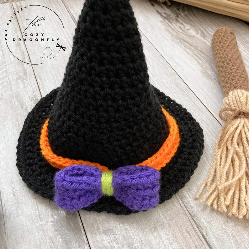 May include: A black crochet witch hat with an orange band and a purple bow. A small crochet broom is next to the hat. The hat has the text "The Cozy Dragonfly" on the left side.