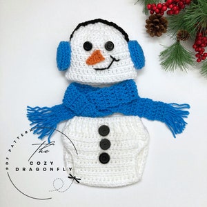 May include: A white crocheted snowman outfit for a baby, including a hat with ear flaps, a scarf, and a diaper cover with black buttons. The hat has black eyes and an orange nose.