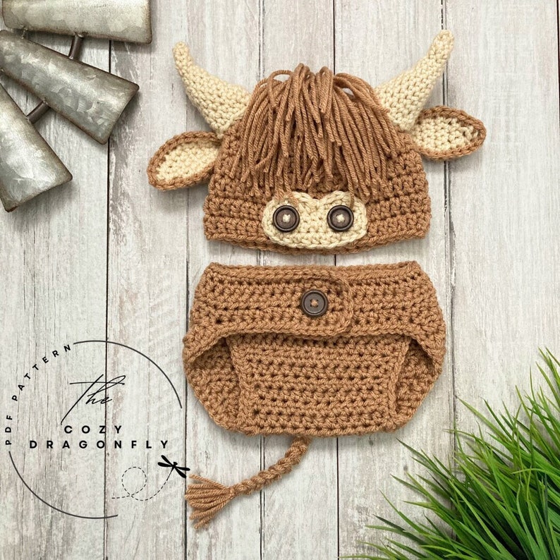 May include: A crocheted baby outfit in the shape of a Highland cow. The outfit includes a hat with ears and a diaper cover with a tail. The hat and diaper cover are brown with cream accents. The hat has two brown buttons for eyes.