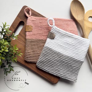 Peut inclure: Deux maniques faites à la main au crochet, roses et blanches, avec des accents marron et gris. Chaque manique a une boucle pour suspendre et une petite étiquette. Les maniques sont sur une planche à découper en bois avec une plante et des cuillères en bois.