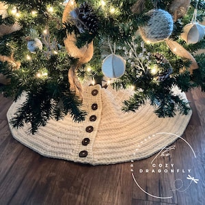 May include: A white crocheted Christmas tree skirt with brown buttons down the centre. The skirt is on a wooden floor and the text "The Cozy Dragonfly" is visible in the bottom right corner.