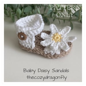 K&ouml;nnte beinhalten: H&auml;kelmuster f&uuml;r Babysandalen mit einem G&auml;nsebl&uuml;mchen-Bl&uuml;tenmuster. Die Sandalen sind wei&szlig; mit einer braunen Sohle und einem Holzknopf. Das Muster hei&szlig;t "Baby Daisy Sandals" und ist von "thecozydragonfly".