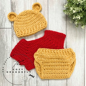 May include: A crocheted baby outfit in red and yellow. The outfit includes a hat, a shirt, and a diaper cover. The hat has bear ears.