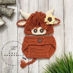May include: Crocheted baby hat and diaper cover set in brown and cream colors, featuring a Highland cow design with a sunflower on the hat. The set includes a tail.