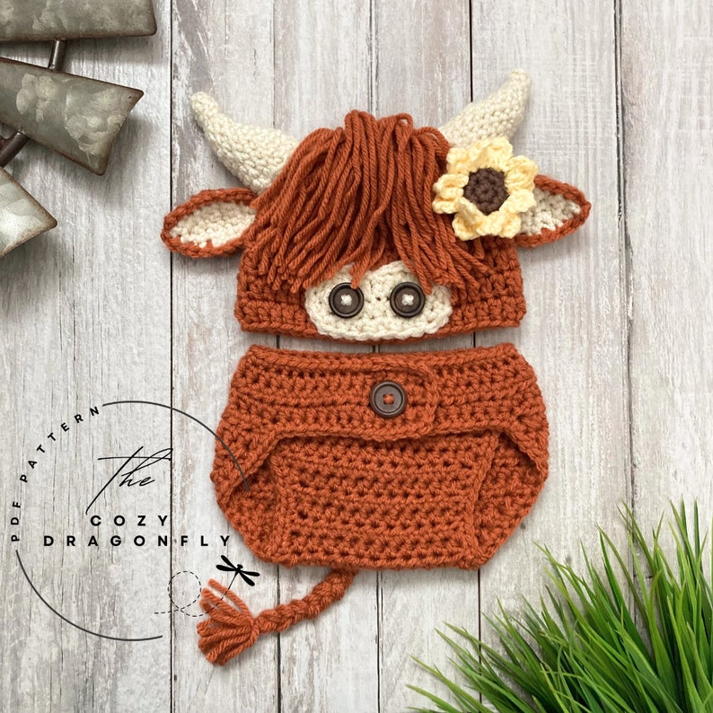 May include: Crocheted baby hat and diaper cover set in brown and cream colors, featuring a Highland cow design with a sunflower on the hat. The set includes a tail.