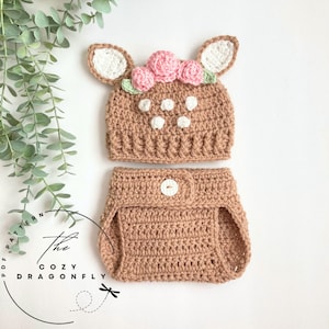 May include: A crochet baby outfit with a deer hat and diaper cover. The hat has brown and white spots, pink flowers, and white ears. The diaper cover is brown and has a white button.