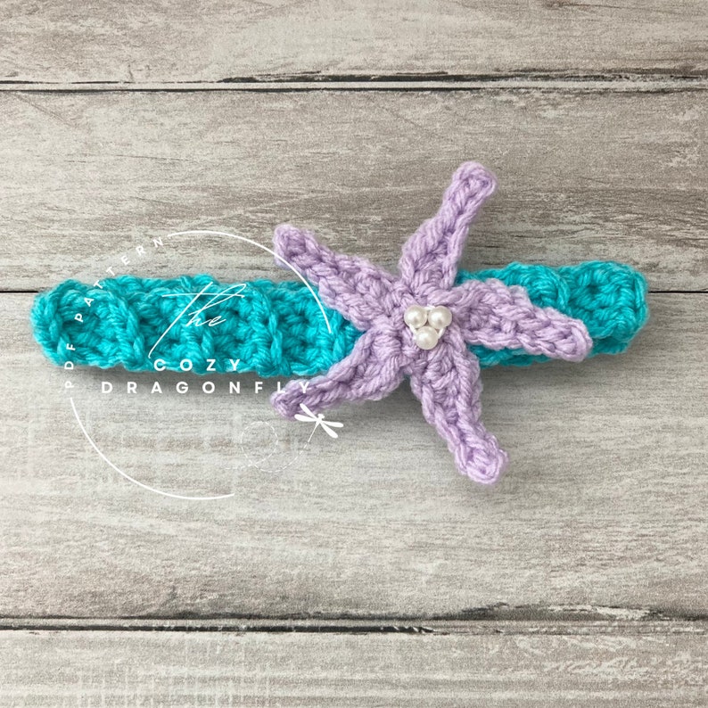May include: A purple crocheted starfish with white pearl beads attached to the center. The starfish is on top of a turquoise crocheted headband.