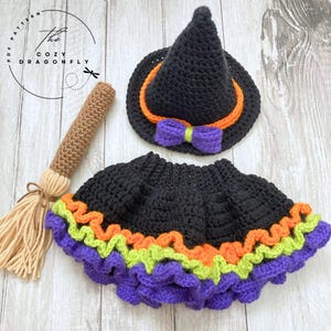 May include: A crochet Halloween costume set featuring a black witch hat with orange trim and a purple bow, a black skirt with purple, orange, and green ruffles, and a brown broom. The Cozy Dragonfly logo is in the upper left corner.