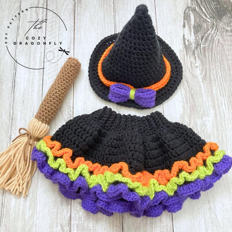 May include: A crochet Halloween costume set featuring a black witch hat with orange trim and a purple bow, a black skirt with purple, orange, and green ruffles, and a brown broom. The Cozy Dragonfly logo is in the upper left corner.