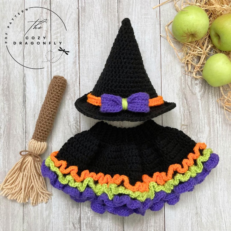 May include: A black crochet witch hat and skirt set with orange, green, and purple trim. A brown crochet broom is also included. The hat has an orange band and a purple bow. The text "The Cozy Dragonfly" is in the upper left corner.