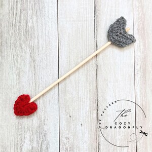 May include: A gray and red crocheted heart arrow with a wooden stick. The arrow is on a white wooden background. The text "The Cozy Dragonfly PDF Pattern" is in a circle on the background.