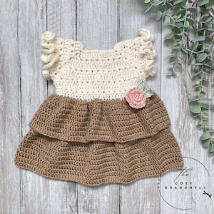 May include: A handmade crochet dress in cream and brown, featuring a ruffled bodice and a tiered skirt. A pink flower with green leaves is attached to the front. The dress is designed for a baby or toddler.