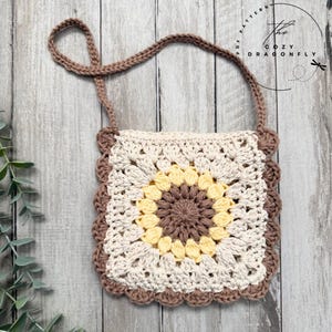 May include: A handmade crochet bag with a sunflower design. The bag features a square shape with a brown strap and scalloped edges. The center of the bag is brown, surrounded by yellow and cream-colored crochet work. The text "The Cozy Dragonfly" is visible.