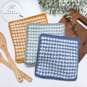 May include: Three square crochet dishcloths featuring a textured stitch pattern in alternating white and solid colours. The set includes one mustard yellow, one sage green, and one dusty blue cloth, each with a consistent white woven design.
