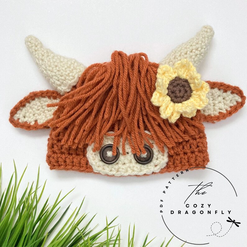 May include: A crocheted highland cow hat with a brown and white color scheme. The hat has a sunflower on the side and button eyes. The hat is sitting on a bed of green grass.