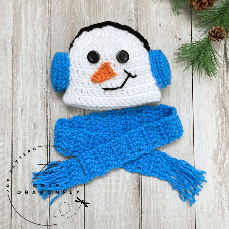 May include: A white crocheted snowman hat with black eyes, an orange nose, and a black mouth. The hat has blue ear flaps and is paired with a blue crocheted scarf with a fringe.