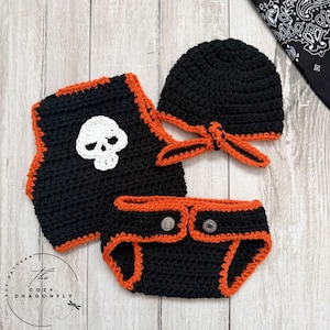 May include: A black and orange crochet baby outfit set, including a hat, diaper cover, and vest. The vest features a white skull applique. The set is trimmed in orange and has silver button closures on the diaper cover.