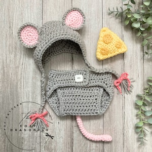 Könnte beinhalten: Ein gehäkeltes Maus-Kostüm für ein Baby, bestehend aus einer grauen Mütze mit rosa Ohren, einer grauen Windelhülle mit Knopf und einem rosa Schwanz. Das Kostüm enthält auch eine gelbe gehäkelte Käse-Requisite.