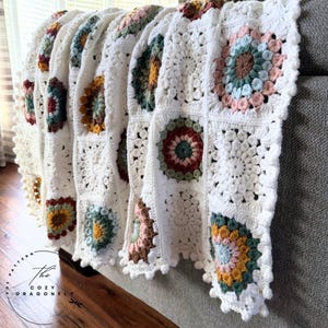May include: A white crochet blanket with colourful floral squares. The blanket features squares with a central flower design in shades of pink, blue, green, and brown. The blanket is draped over a grey surface, with the text "The Cozy Dragonfly" visible.