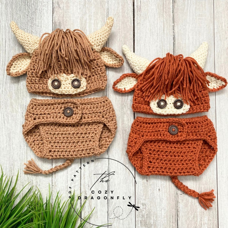 May include: Two crocheted baby outfits in the shape of a Highland cow. The outfits are brown and orange and feature a hat, diaper cover, and tail. The outfits are on a wooden background.
