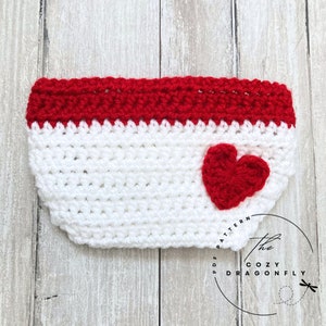 May include: White crocheted cup cozy with a red crocheted heart and red crocheted trim. The cozy has the text "The Cozy Dragonfly" and "PDF Pattern" on the side.