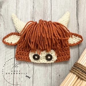 May include: A crocheted hat shaped like a Highland cow with brown and white yarn. The hat has a fringe of brown yarn and two black buttons for eyes.
