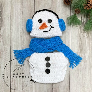 May include: A white crocheted snowman costume for a baby, featuring a hat with ear flaps, a scarf, and a diaper cover with three black buttons. The hat has black eyes, an orange nose, and a black mouth.