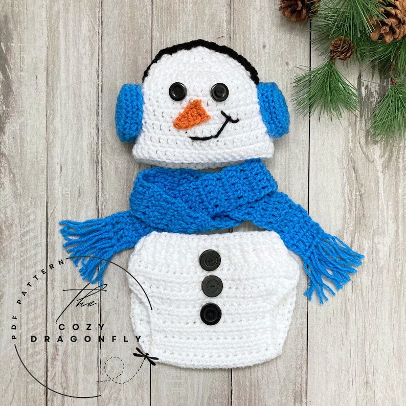 May include: A white crocheted snowman costume for a baby, featuring a hat with ear flaps, a scarf, and a diaper cover with three black buttons. The hat has black eyes, an orange nose, and a black mouth.